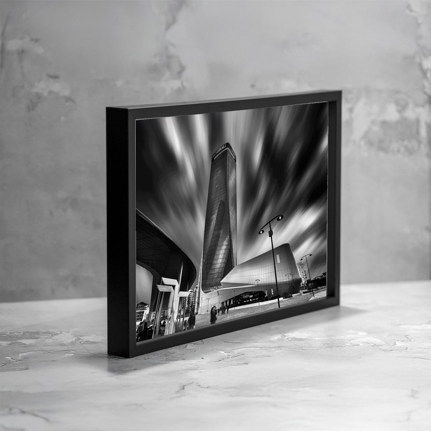 Fotografia fine art in bianco e nero incorniciata di architettura contemporanea a Milano, con il grattacielo Generali ripreso dal basso e una lunga esposizione che genera un cielo dinamico e mosso, presentata in una vista angolata come esempio di ambientazione.
Black and white fine art photograph shown framed of contemporary architecture in Milan, featuring the Generali tower shot from a low angle, with long exposure creating a dynamic, streaked sky, presented in an angled view as an example of display.