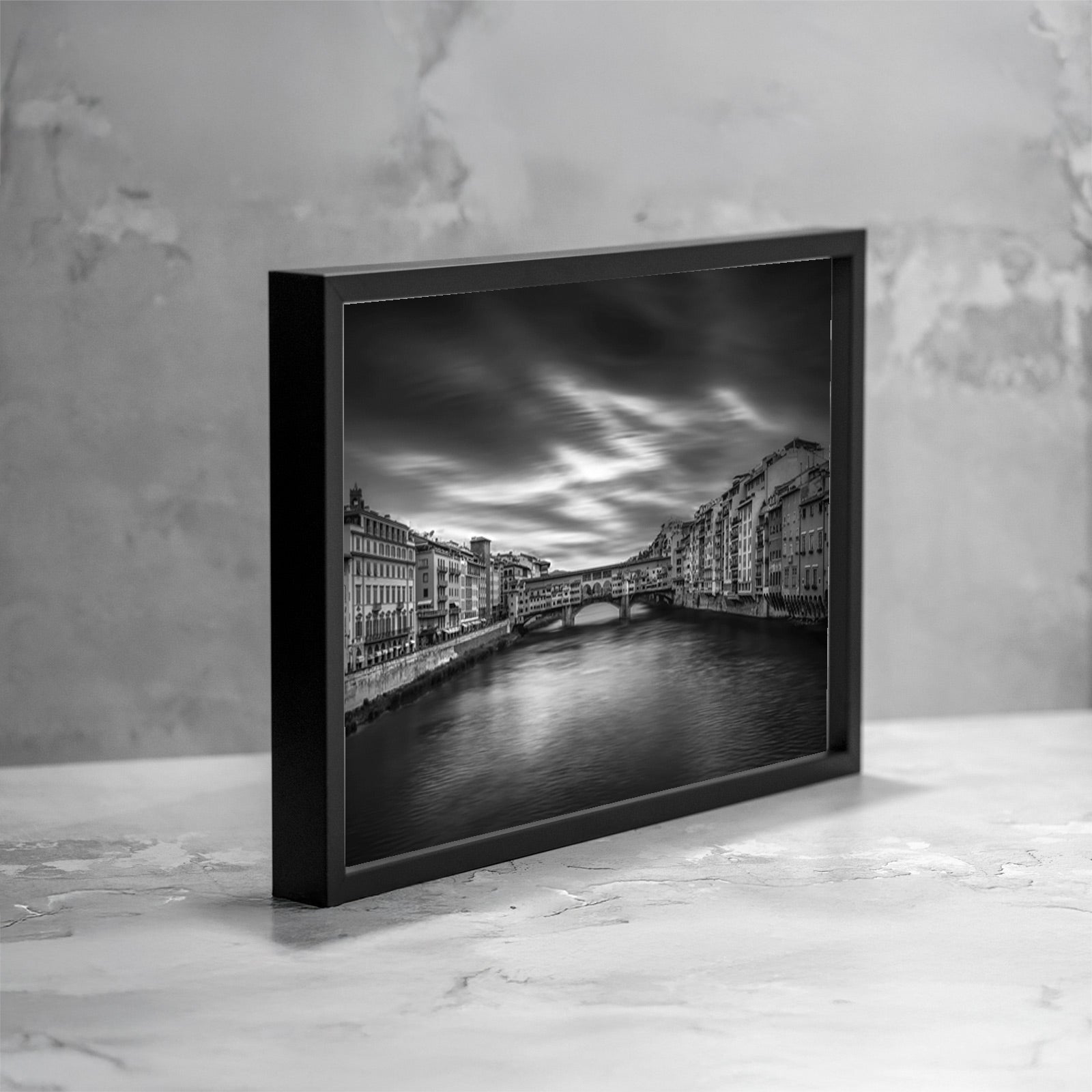 Fotografia fine art in bianco e nero incorniciata, raffigurante Ponte Vecchio a Firenze visto frontalmente sul fiume Arno, con lunga esposizione che rende l’acqua e il cielo morbidi e sfumati, presentata in una vista angolata come esempio di ambientazione.
Black and white fine art photograph shown framed, depicting Ponte Vecchio in Florence viewed frontally over the Arno River, with long exposure creating smooth water and soft clouds, presented in an angled view as an example of display.