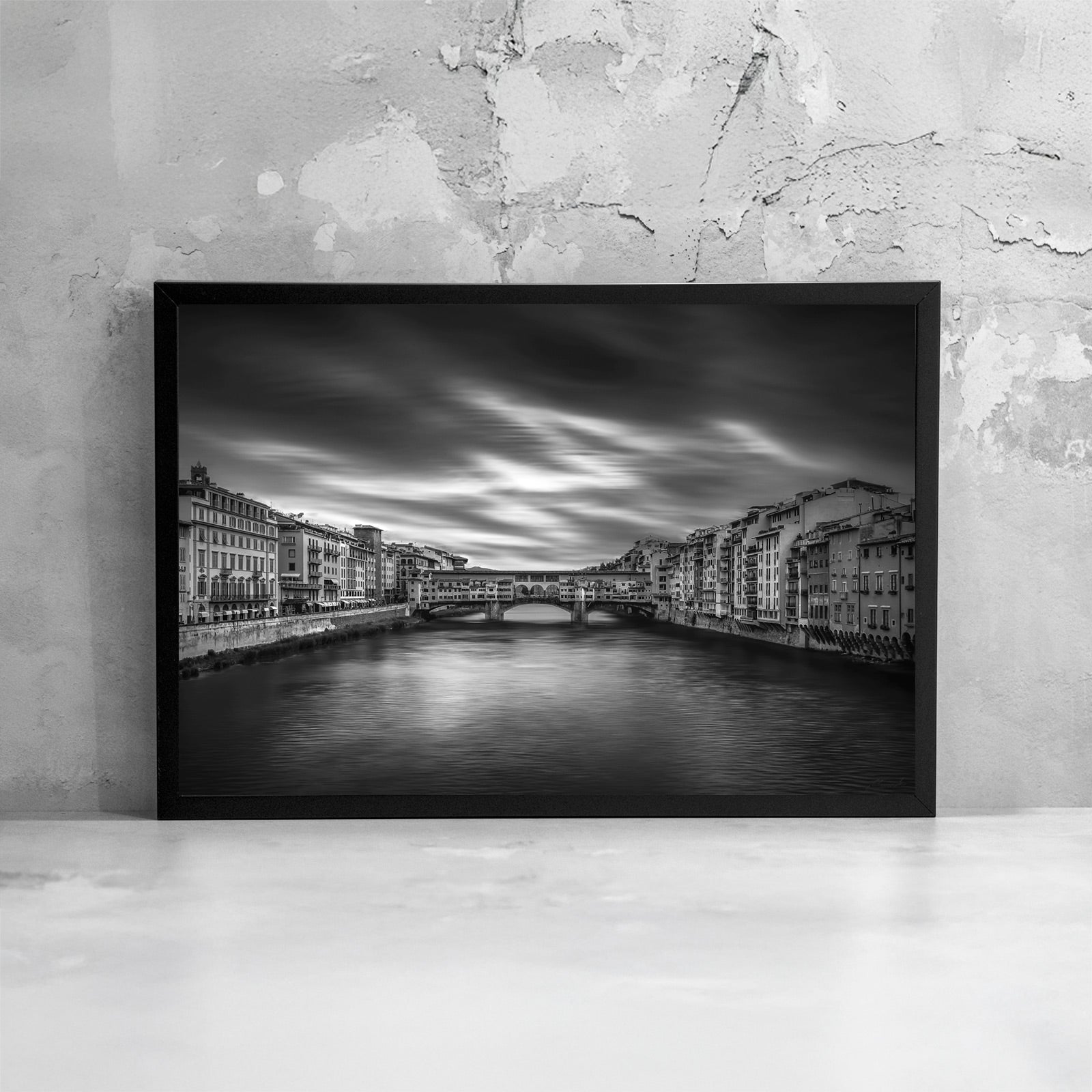 Fotografia fine art in bianco e nero incorniciata di Ponte Vecchio a Firenze, ripreso frontalmente sul fiume Arno con lunga esposizione, presentata come dettaglio di stampa e cornice.
Black and white fine art photograph shown framed of Ponte Vecchio in Florence viewed over the Arno River with long exposure, presented as a print and frame detail.
