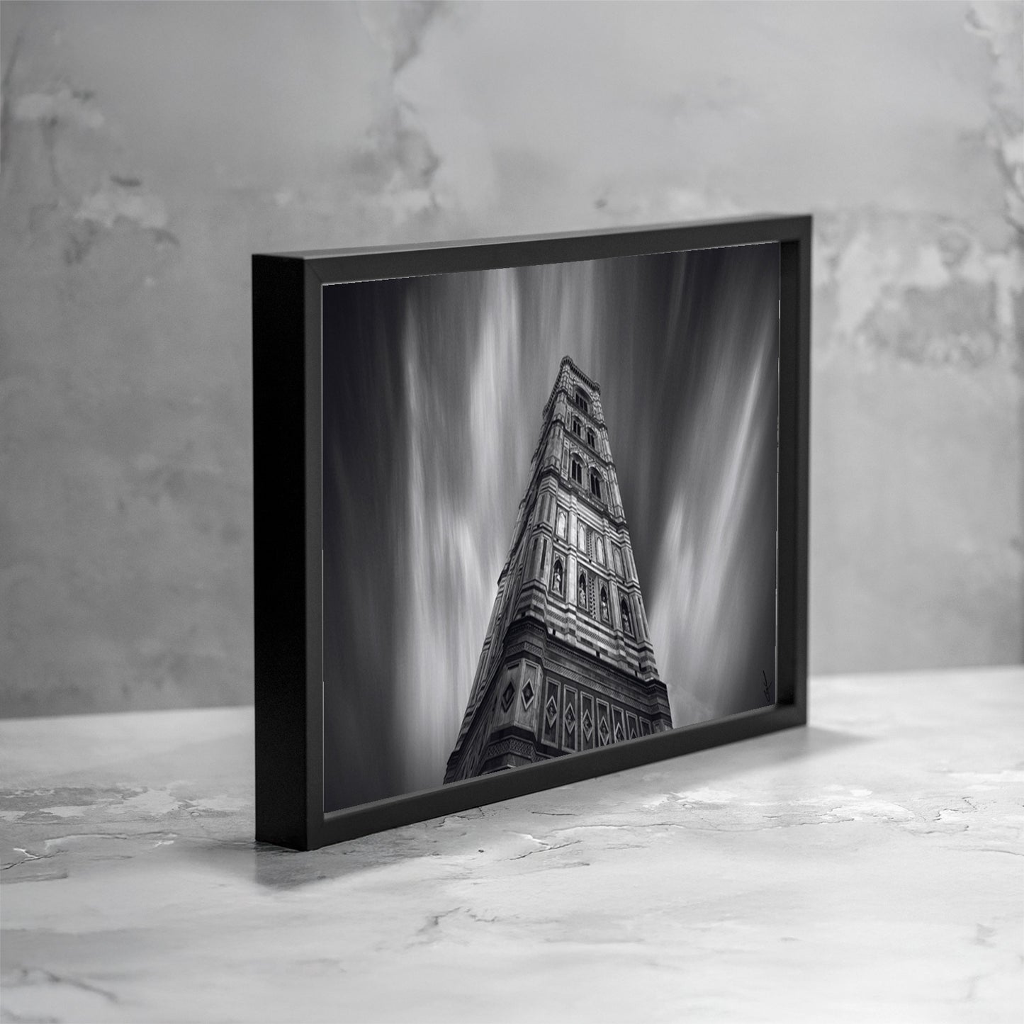 Fotografia fine art in bianco e nero incorniciata del Campanile di Giotto a Firenze, ripreso dal basso con una lunga esposizione che crea scie verticali nel cielo, presentata in una vista angolata come esempio di ambientazione.
Black and white fine art photograph shown framed of Giotto’s Bell Tower in Florence, shot from a low angle with long exposure creating vertical light streaks in the sky, presented in an angled view as an example of display.