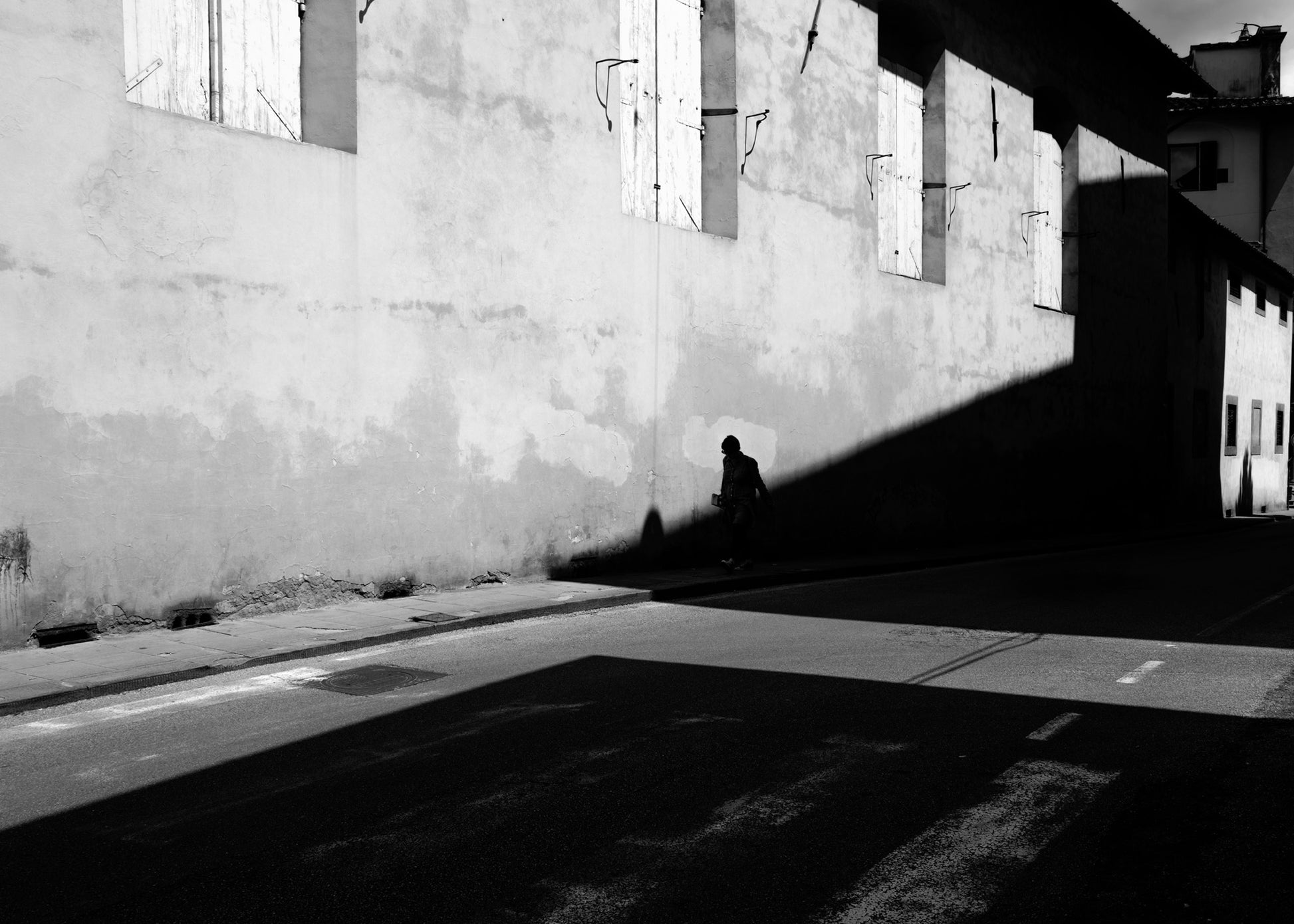 Una figura solitaria attraversa una linea di confine tracciata dalla luce e dall’ombra lungo una parete urbana, generando una composizione essenziale fatta di silenzio, spazio e tensione visiva.
A solitary figure crosses a boundary drawn by light and shadow along an urban wall, creating an essential composition shaped by silence, space, and visual tension.