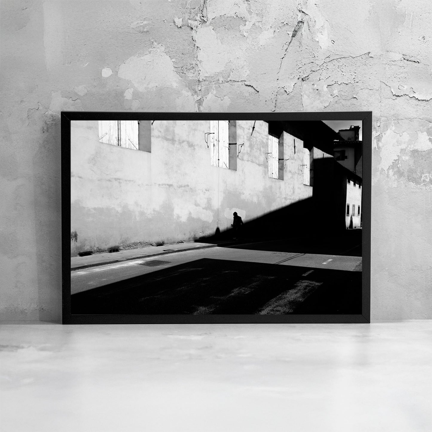 La fotografia incorniciata enfatizza il contrasto tra luce e oscurità, trasformando la scena urbana in un’immagine minimale dove la presenza umana diventa misura dello spazio.
The framed photograph emphasizes the contrast between brightness and darkness, transforming the urban scene into a minimal image where human presence becomes the measure of space.