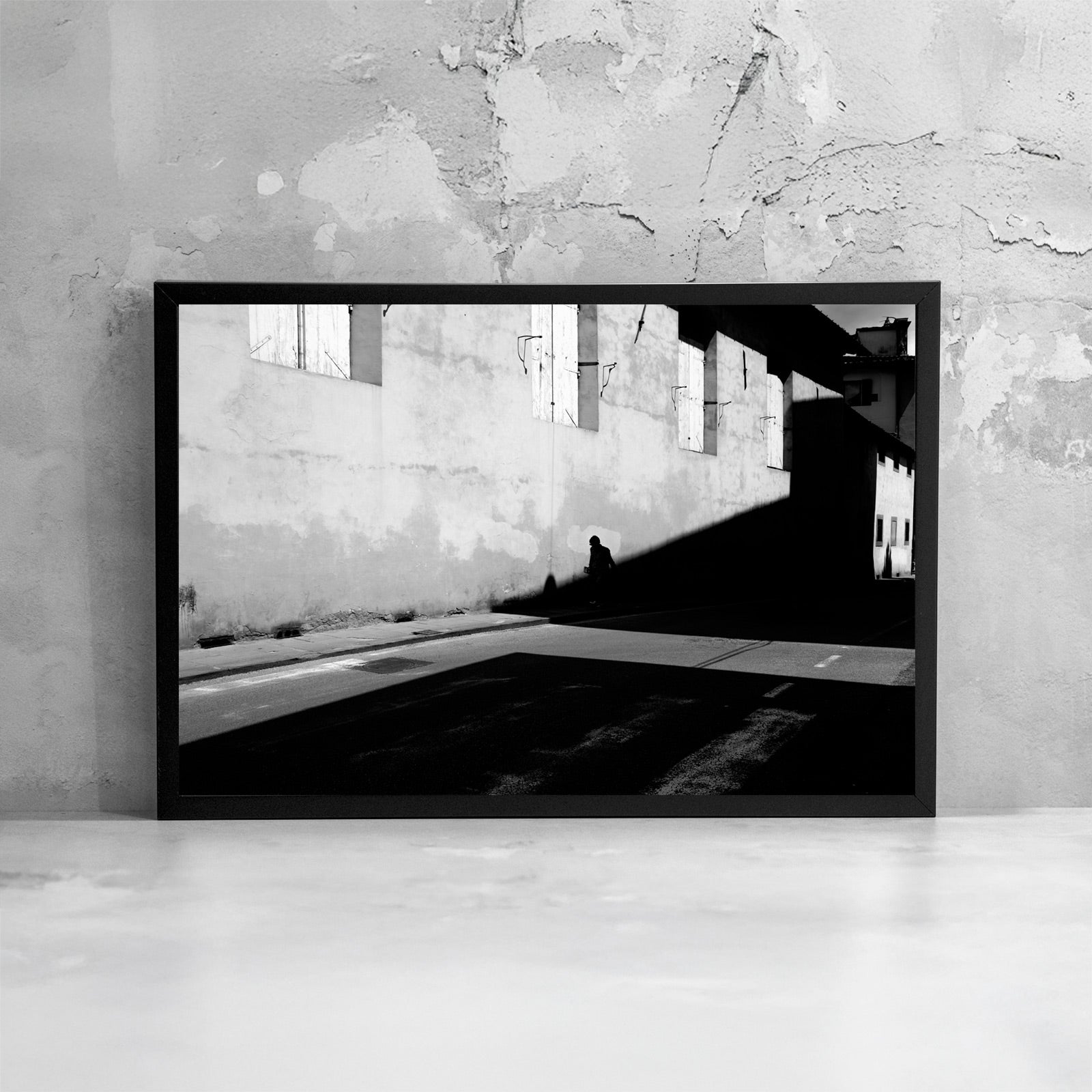 La fotografia incorniciata enfatizza il contrasto tra luce e oscurità, trasformando la scena urbana in un’immagine minimale dove la presenza umana diventa misura dello spazio.
The framed photograph emphasizes the contrast between brightness and darkness, transforming the urban scene into a minimal image where human presence becomes the measure of space.