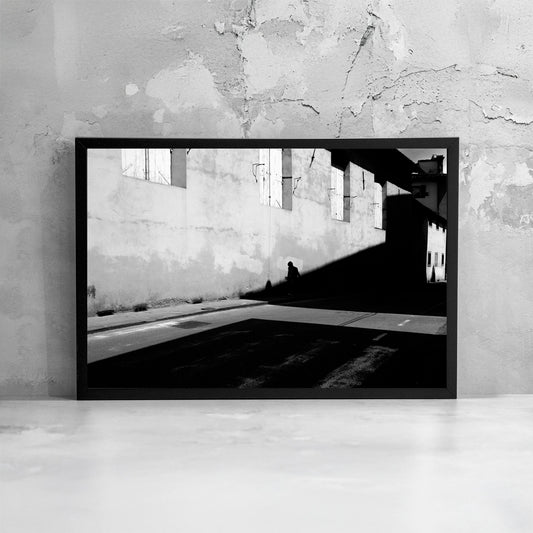 La fotografia incorniciata enfatizza il contrasto tra luce e oscurità, trasformando la scena urbana in un’immagine minimale dove la presenza umana diventa misura dello spazio.
The framed photograph emphasizes the contrast between brightness and darkness, transforming the urban scene into a minimal image where human presence becomes the measure of space.